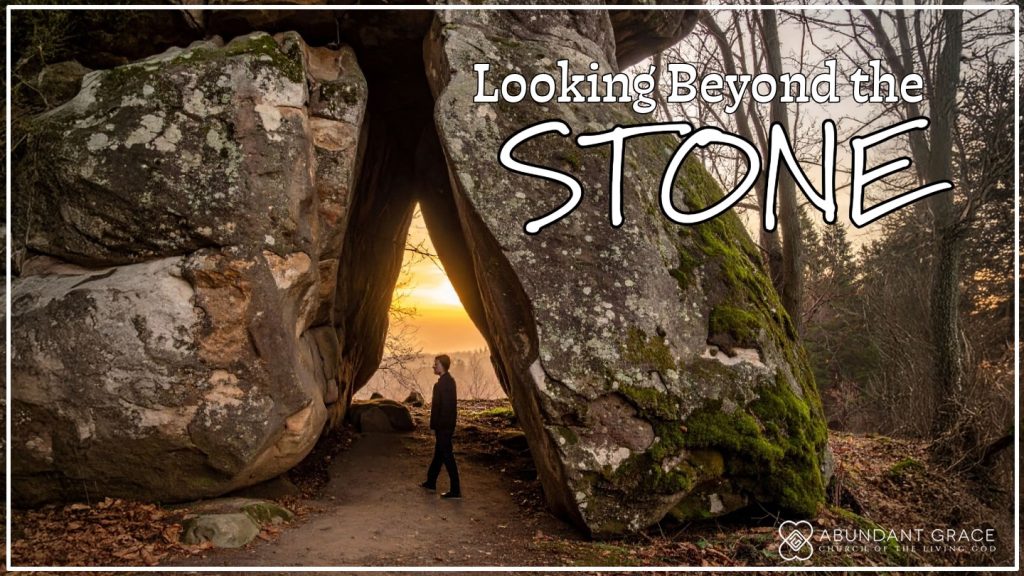 A person standing in the woods, gazing at a massive stone that blocks the entrance of a cave.